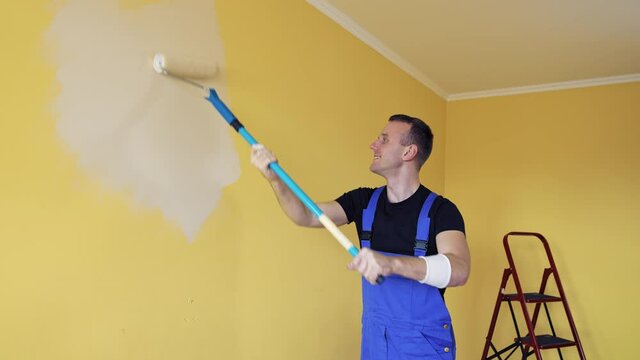 Room improvement with a new color. Professional worker in blue overalls painting wall with a paint roller on a long handle on. Craftsman working.