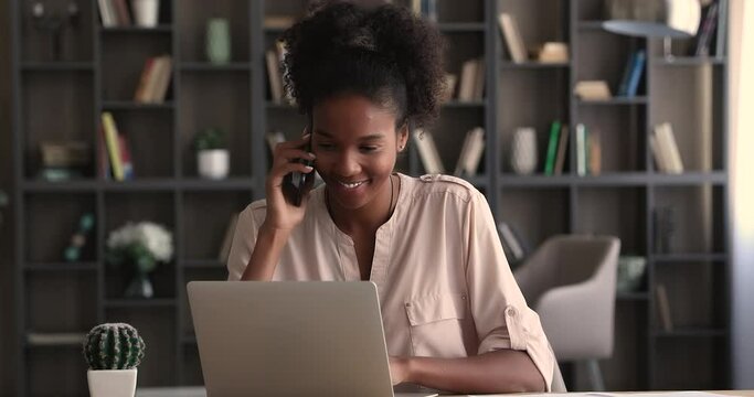 Focused millennial african american business lady working on computer, giving consultation in cellphone conversation to client or discussing project issues with colleagues distantly, multitask concept