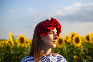 Young woman wearing a wreath of red flowers looks towards the sunset on a sunflower field