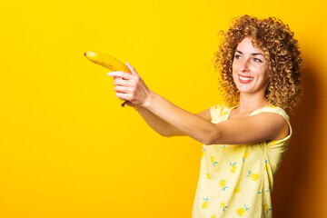 smiling curly young woman holding a banana pistol on a yellow background