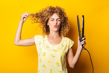 grimaced young woman straightens curly hair with a hair straightener on a yellow background
