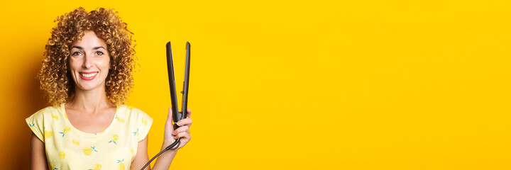 smiling young woman with curly hair holding hair straightener on yellow background. Banner.