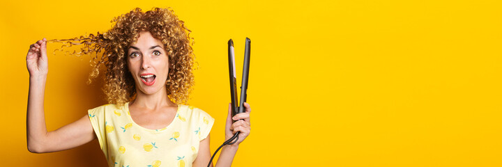 surprised young woman with curly hair with hair straightener on yellow background. Banner.