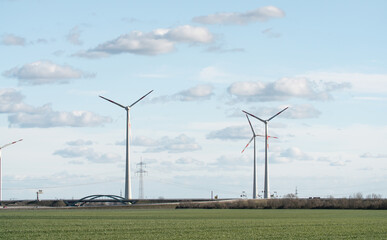 Fototapeta premium Some wind turbines on a field, clouds and a blue sky. A bridge in the background.