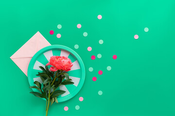 Green stripy metal tray with single pink peony flower over paper envelope on green background with...
