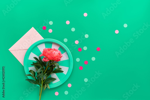 Green stripy metal tray with single pink peony flower over paper envelope on green background with text space. Trendy casual natural eco friendly flat lay. Summer birthday or Mother's day greeting