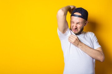 Young man in a cap and T-shirt sniffs his armpits on a yellow background. Sweat concept, sweat stains - obrazy, fototapety, plakaty