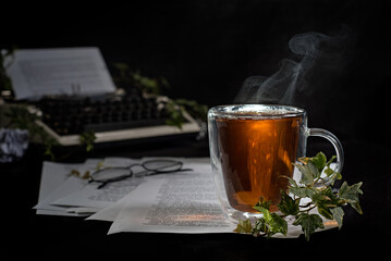 A cup of hot tea with steam stands on sheets of paper with text. Against the background of a vintage typewriter, black-framed glasses and ivy branches with green leaves.