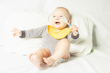 Little baby in a yellow children's apron with food residue and a spoon in his hand is sitting on a...