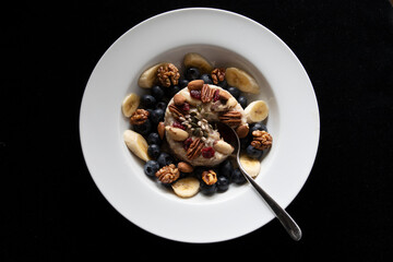 WHITE DISH ORGANIC PORRIDGE BLUEBERRIES BANANA NUTS SEEDS SPOON ON BLACK 