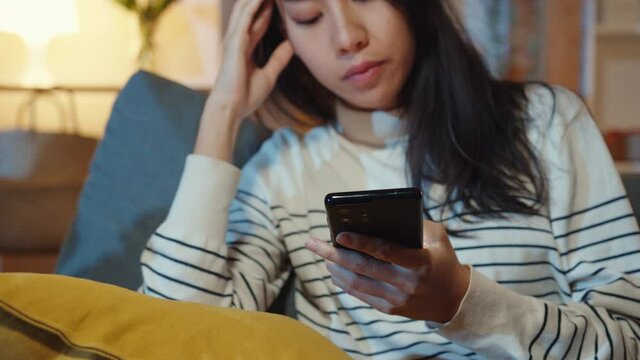 Thoughtful Asia lady holding phone feeling sad waiting for call sit at sofa in living room at house night feel lonely, Sad depressed teenager spend time alone, Social distance, Coronavirus quarantine.