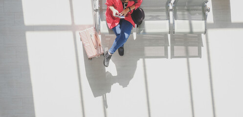 Top view young hipster female traveler with suitcase using smartphone during departure time at public terminal station with copy space. Travel alone minimal background concept.