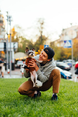 Cheerful smiling man in casual attire is feeling happy when holding his adorable dog - york terrier, sitting on a lawn near the city street. Vertical shot.