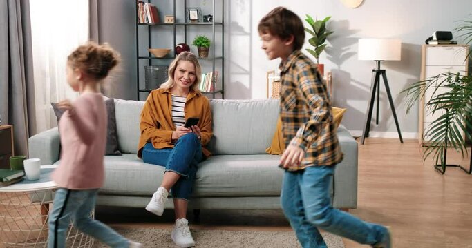 Cheerful adorable Caucasian small children boy and girl running and playing in apartment while their mom sitting on sofa at home typing on smartphone browsing online and smiling, family concept