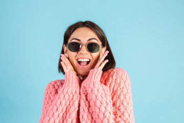 Young brunette in warm pink sweater isolated on blue background happy look to camera shocked amazed surprised open mouth