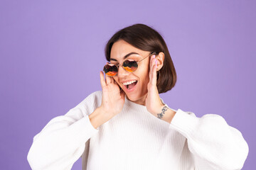 Young brunette in white casual sweater isolated on purple background wearing sunglasses happy smile positive moving