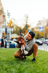 Vertical photo of a joyful funny young male dog-owner holding his cute doggy - york terrier, sitting on a green lawn near the busy city street.