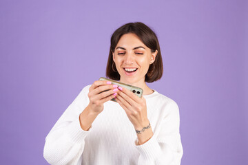 Young brunette in white casual sweater isolated on purple background happy overjoyed playing games on mobile phone