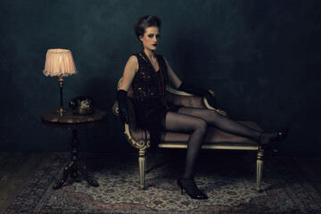 Seductive young woman in retro boudoir attire with necklaces on an antique sofa next to a coffee table with a table lamp and a telephone.