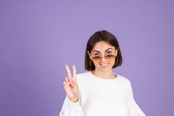 Young brunette in white casual sweater isolated on purple background smile to camera do peaceful gesture victory sign