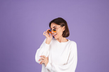 Young brunette in white casual sweater isolated on purple background in sunglasses happy stylish positive smiling