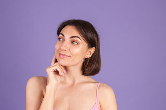 Young brunette in top isolated on purple background looks to the side thoughtfully with a sweet smile