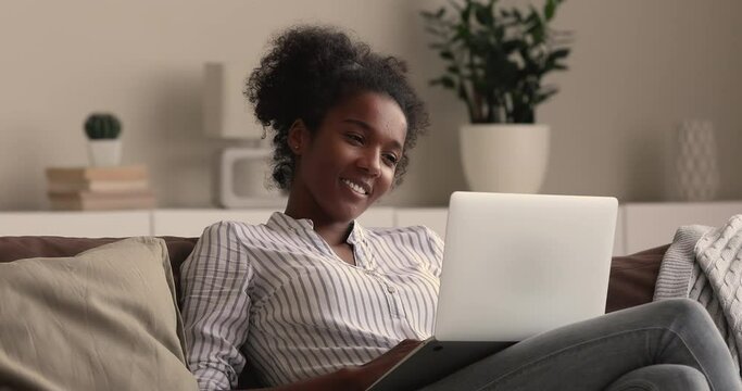 Relaxed millennial smiling african mixed race woman looking at laptop screen, watching funny movie film or pleasant video photo content in social network, shopping in internet store or web surfing.