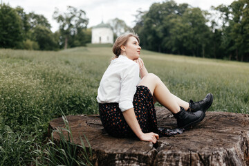 Fototapeta premium A young stylish girl sits on a stump against a background of grass and looks to the side. Place for your text.