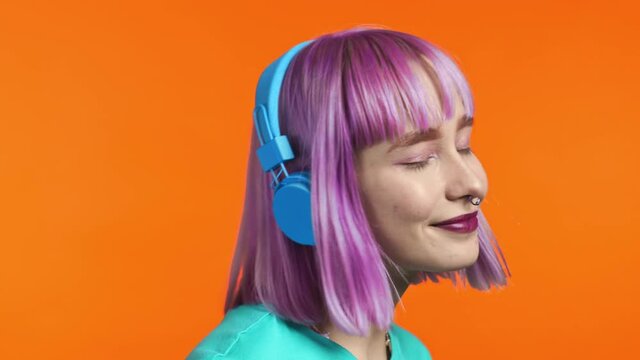 Attractive modern woman dancing with headphones on orange studio background. Cute girls with violet dyed hair portrait. Music, radio, happiness concept.