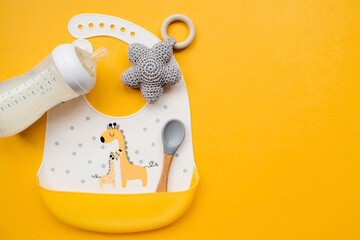Serving baby food. Colorful silicone bib with giraffes, spoon and milk bottle on a trendy yellow...