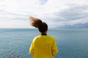 Fototapeta premium Back photography of young woman wearing yellow long sleeve stands in front of the sea under blue sky and white clouds.