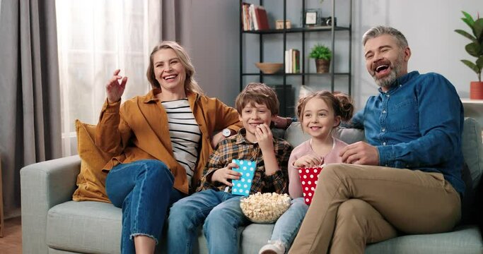 Caucasian joyful happy Caucasian family resting at home watching TV together sitting on couch with popcorn and laughing. Mom and dad with small son and daughter eating popcorn watching comedy movie