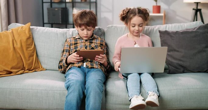 Cute Caucasian little children sitting on sofa in room using digital gadgets browsing online. Small cute girl surfing internet on laptop while kid boy playing game on tablet device, family concept