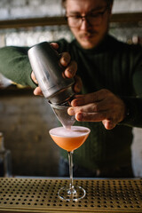 Bartender pours whiskey sour cocktail from metal shaker into glass through the sieve.