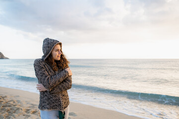 Young woman on the beach standing against the sunset with delight expression. Warming embrace own body.