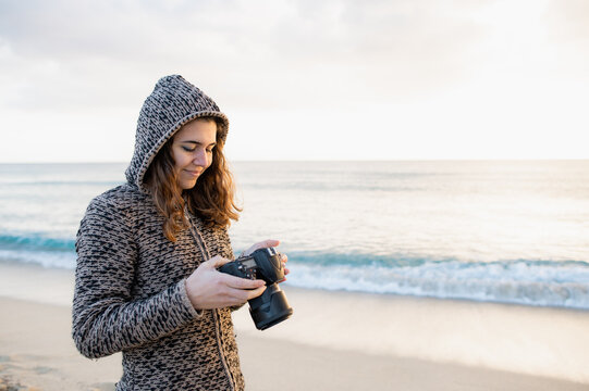 Woman in casual clothing with hood standing in front a sandy beach during sunset and holding and setting camera to take photos of the landscape.