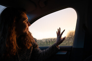 Portrait of young woman inside car while raining. Smiley face looking the sunset out the window mirror with drops of rain.