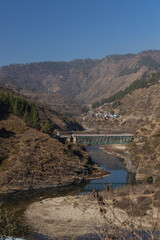 Fototapeta na wymiar View of a river flowing through a narrow valley with rocky mountains on the sides and a bridge on top of it in the Kumaun region of UttaraKhand India on 12 January 2021 