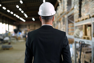 Back view of a boss in a suit and white helmet walking through a work warehouse
