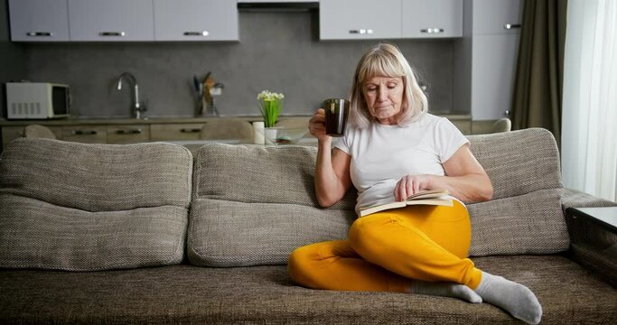 Senior woman or grandmother relaxing on a sofa reading a book and enjoying a mug of hot tea or coffee with copyspace alongside