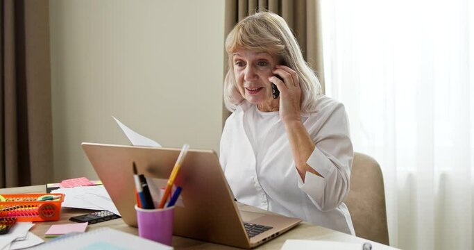 Senior Woman Working From Home During The Covid-19 Pandemic Discussing Paperwork With A Client Or Colleague On The Phone As She Sits At Her Laptop Computer