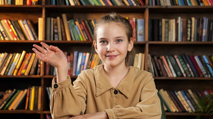 Schoolgirl in brown jacket waves hand talks and smiles to camera sitting at online lesson at home against coloured books on shelves closeup