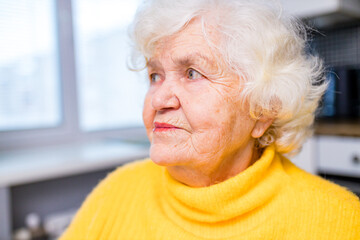 Thoughtful senior lady sitting at home with her fingers to her chin in living room