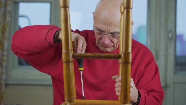Elderly man in glasses and with screwdriver in hands repairs wooden stool