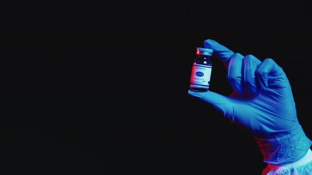 Covid-19 vaccine. Stop pandemic. Disease prevention. Dose vial in doctor hand in blue protective glove in red neon light isolated on black empty space background.