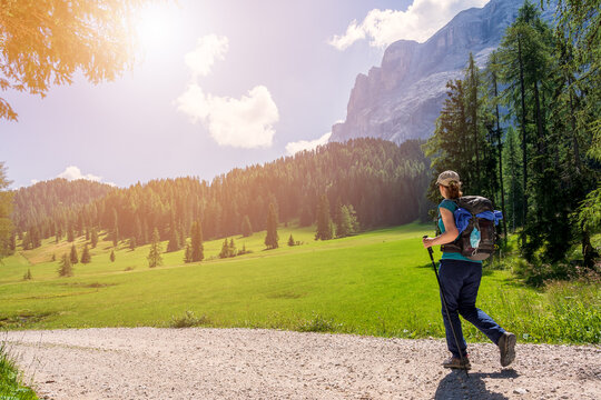Summer: young woman with a hat, trekking poles, a backpack and a face mask attached to it, hiking on a trail through green meadows in the Italian Alps. Dolomite peaks are visible in the background - Powered by Adobe