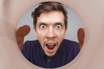 Man looking inside cup or rounded tube. Close-up shot of shocked young man with round eyes and open mouth.