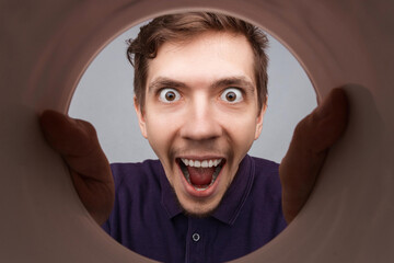 Man looking inside cup or rounded tube with hands on it. Close-up shot of happy shocked young man with round eyes and open mouth  smile.