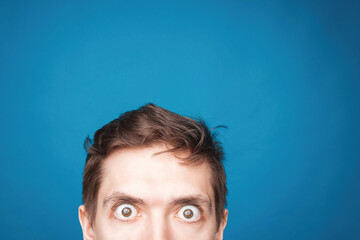 Man's eyes looking up on the light background. Empty place for a text or object. Close-up shot of shocked young man with round eyes, top half head. Eyes looking on front