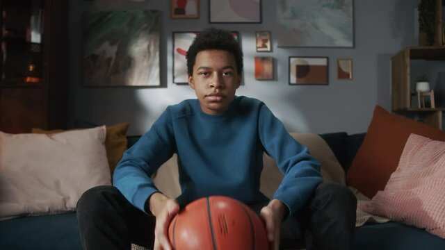 Serious black man teenager holding basketball ball and sitting on sofa in living room, young teen athlete, sport portrait at home, high school kid preparing for watching professional game on tv.